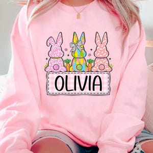 May include: Pink sweatshirt with a graphic of three Easter bunnies with different patterns. The bunnies are sitting on a checkered banner with the name "Olivia" written in black.