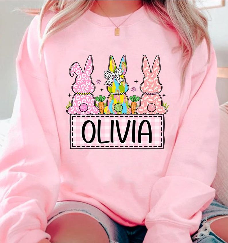May include: Pink sweatshirt with a graphic of three Easter bunnies with different patterns. The bunnies are sitting on a checkered banner with the name "Olivia" written in black.