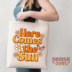 May include: A white tote bag with the text "Here Comes the Sun" in orange and yellow letters. The text is surrounded by small white daisies and a butterfly. The text "DESIGN SOUL" is in the bottom right corner.