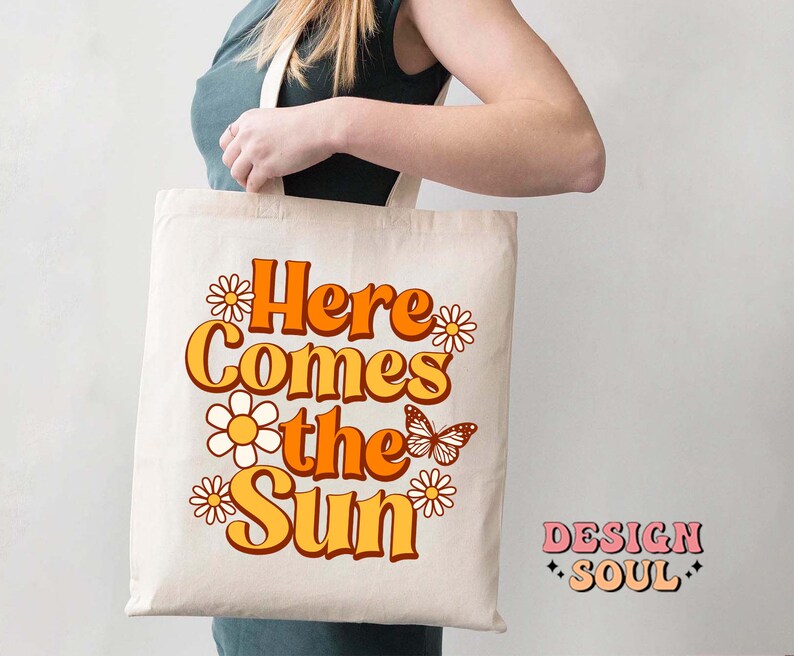 May include: A white tote bag with the text "Here Comes the Sun" in orange and yellow letters. The text is surrounded by small white daisies and a butterfly. The text "DESIGN SOUL" is in the bottom right corner.