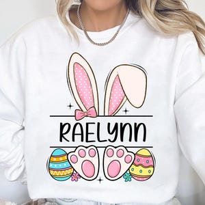 May include: White sweatshirt with a pink bunny ears design, the name "RAELynn" in black text, and two pastel Easter eggs with paw prints.