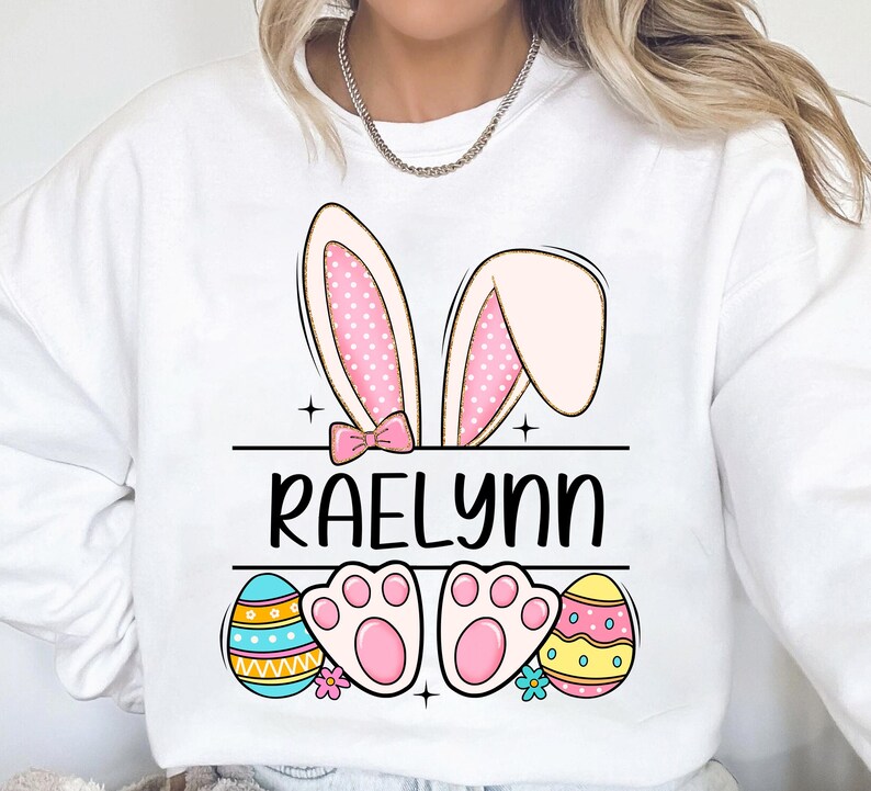 May include: White sweatshirt with a pink bunny ears design, the name "RAELynn" in black text, and two pastel Easter eggs with paw prints.
