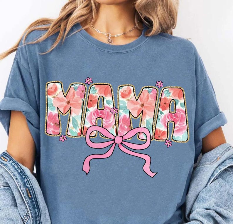 May include: A blue t-shirt with the word "MAMA" in pink and white floral letters with a pink bow underneath. The letters have a gold glitter outline.