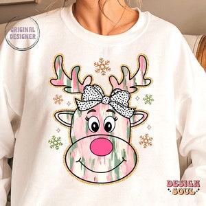 May include: White sweatshirt with a pink and green reindeer design. The reindeer has a black and white polka dot bow and a pink nose. The design is surrounded by gold glitter snowflakes.