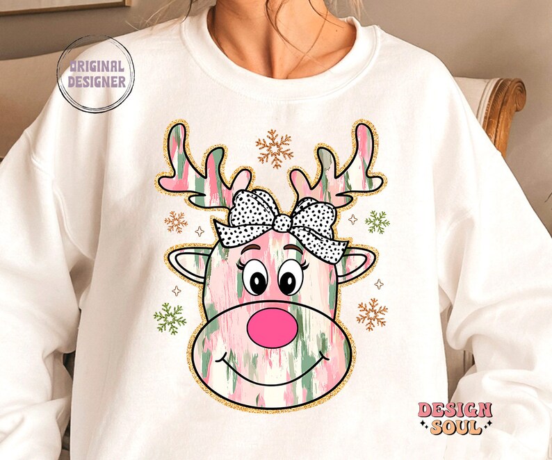 May include: White sweatshirt with a pink and green reindeer design. The reindeer has a black and white polka dot bow and a pink nose. The design is surrounded by gold glitter snowflakes.