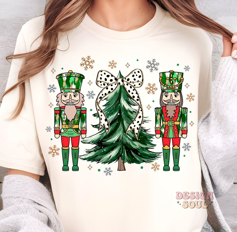May include: A cream-coloured t-shirt with a festive Christmas design. The design features two nutcrackers in green and red uniforms, a green Christmas tree, and a black and white polka dot bow. Snowflakes and the words "Design Soul" are also present.