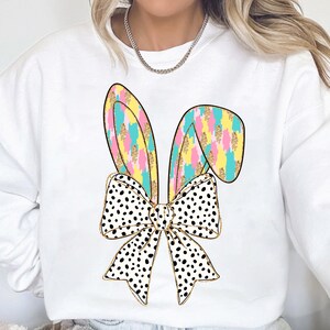 May include: White sweatshirt with a colorful bunny ear bow design. The bow is black and white polka dot with gold glitter accents. The bunny ears are pink, yellow, blue, and green.