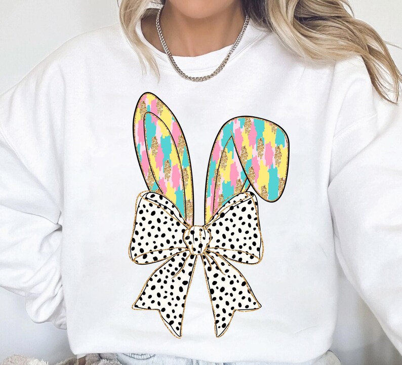May include: White sweatshirt with a colorful bunny ear bow design. The bow is black and white polka dot with gold glitter accents. The bunny ears are pink, yellow, blue, and green.