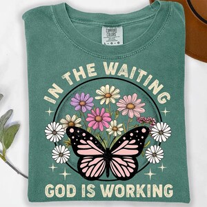 May include: A teal green t-shirt with a floral design and a butterfly. The text "In the waiting, God is working" is printed on the shirt.