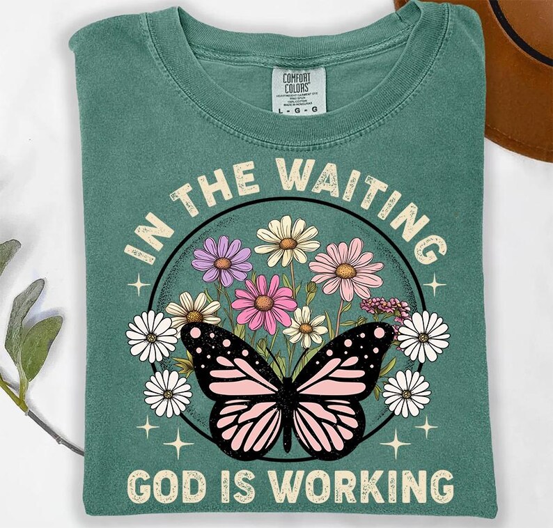 May include: A teal green t-shirt with a floral design and a butterfly. The text "In the waiting, God is working" is printed on the shirt.