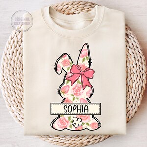 May include: A cream-colored sweatshirt with a floral bunny design and the name "Sophia" printed on a banner. The bunny is outlined in black and has a pink bow.