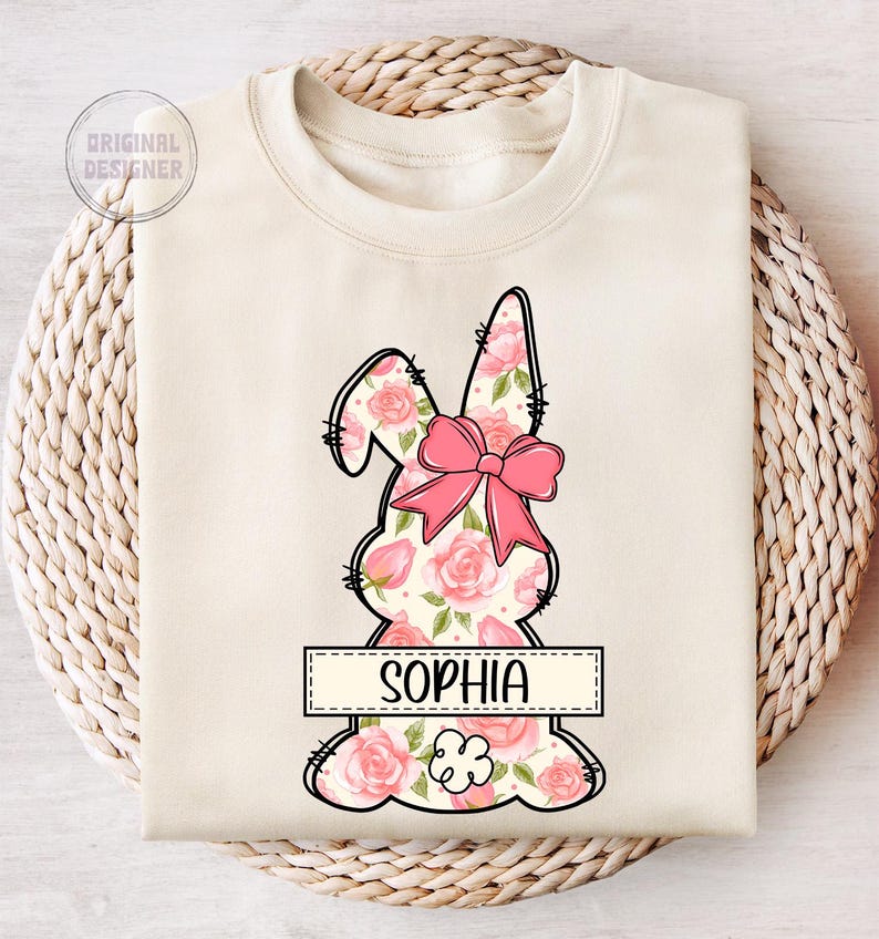 May include: A cream-colored sweatshirt with a floral bunny design and the name "Sophia" printed on a banner. The bunny is outlined in black and has a pink bow.