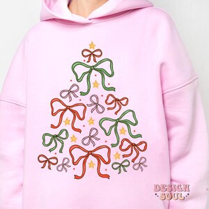May include: Pink hoodie featuring a Christmas tree design made of bows in various colours, including green, red, pink, and yellow. The tree is decorated with gold stars.