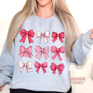 May include: A light gray sweatshirt with a design of nine pink bows of different shapes and sizes. The sweatshirt has a crew neck and long sleeves.
