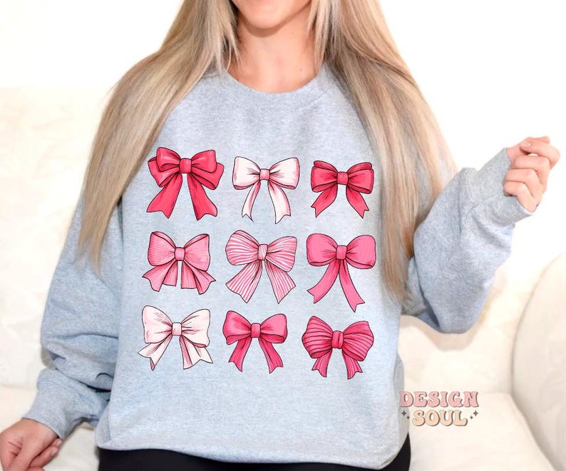 May include: A light gray sweatshirt with a design of nine pink bows of different shapes and sizes. The sweatshirt has a crew neck and long sleeves.