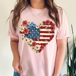 May include: A light pink t-shirt featuring a heart-shaped design of the American flag. The flag design is surrounded by red, white, and blue flowers. The shirt is a casual style.