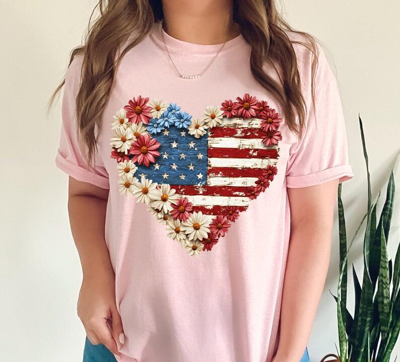 May include: A light pink t-shirt featuring a heart-shaped design of the American flag. The flag design is surrounded by red, white, and blue flowers. The shirt is a casual style.