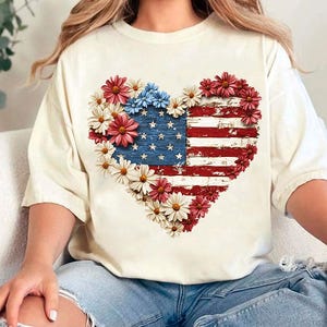May include: Cream-colored t-shirt featuring a heart-shaped design of the American flag, adorned with red, white, and blue flowers. The flag design has a distressed, vintage look, with visible stars and stripes. The shirt is a casual, everyday wear item.