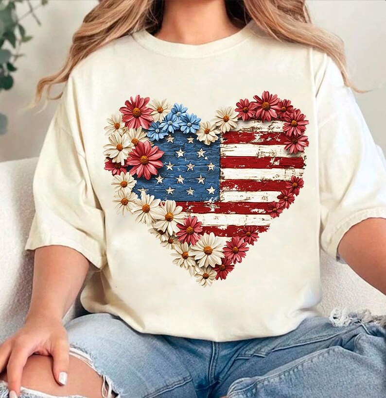 May include: Cream-colored t-shirt featuring a heart-shaped design of the American flag, adorned with red, white, and blue flowers. The flag design has a distressed, vintage look, with visible stars and stripes. The shirt is a casual, everyday wear item.