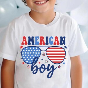 May include: White t-shirt with a red, white, and blue American flag design. The design features sunglasses with the American flag pattern and the text "AMERICAN boy".