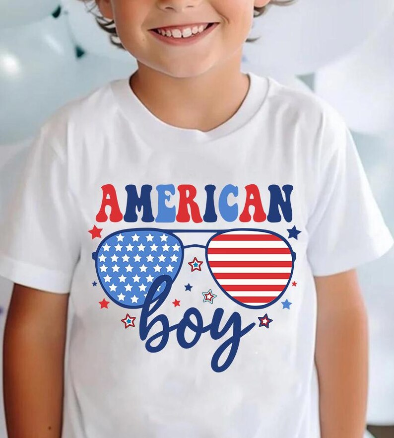 May include: White t-shirt with a red, white, and blue American flag design. The design features sunglasses with the American flag pattern and the text "AMERICAN boy".