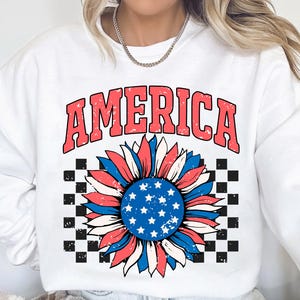 May include: White sweatshirt with a red, white, and blue sunflower graphic and the word "AMERICA" in red letters. The sunflower has a checkered background.