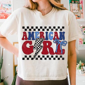 May include: A white t-shirt with a red, white, and blue graphic that says "American Girl". The graphic includes a lightning bolt with a checkered pattern and a blue bow.