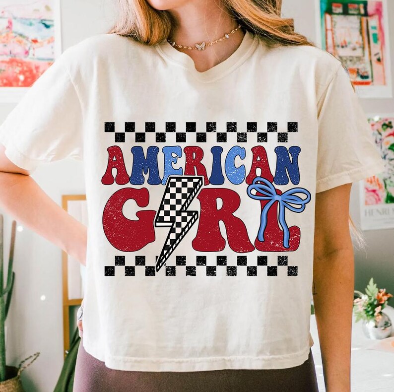 May include: A white t-shirt with a red, white, and blue graphic that says "American Girl". The graphic includes a lightning bolt with a checkered pattern and a blue bow.