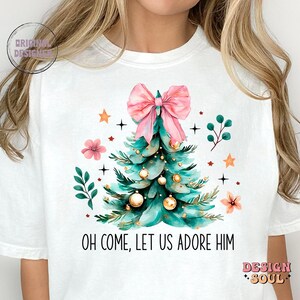 May include: White t-shirt with a Christmas tree design featuring a pink bow, gold ornaments, and green leaves. The text "Oh come, let us adore Him" is printed below the tree.