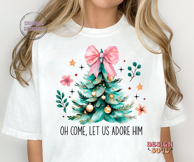 May include: White t-shirt with a Christmas tree design featuring a pink bow, gold ornaments, and green leaves. The text "Oh come, let us adore Him" is printed below the tree.