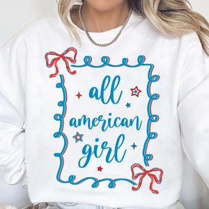 Peut inclure: Sweat-shirt blanc avec un graphique bleu et rouge qui dit "all american girl" &agrave; l'int&eacute;rieur d'un cadre bleu et rouge avec des &eacute;toiles et des n&oelig;uds.