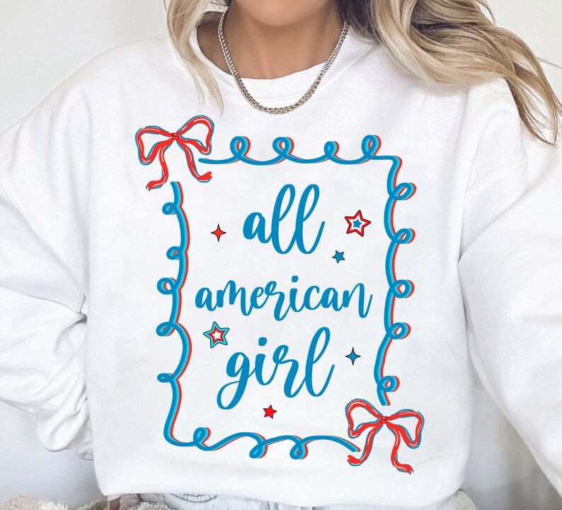 Peut inclure: Sweat-shirt blanc avec un graphique bleu et rouge qui dit "all american girl" &agrave; l'int&eacute;rieur d'un cadre bleu et rouge avec des &eacute;toiles et des n&oelig;uds.