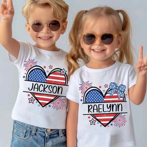 May include: Two children wearing white t-shirts with a patriotic heart design. The heart features the American flag colors and the names "Jackson" and "Raelynn". The shirts are decorated with fireworks and stars.