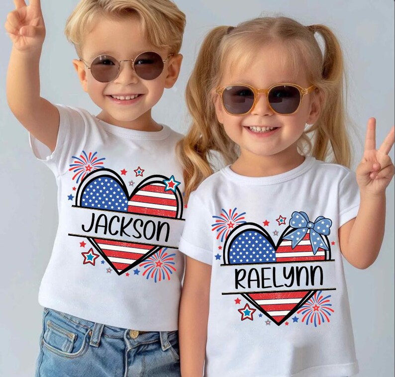 May include: Two children wearing white t-shirts with a patriotic heart design. The heart features the American flag colors and the names "Jackson" and "Raelynn". The shirts are decorated with fireworks and stars.
