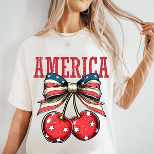 May include: White t-shirt with a red, white, and blue bow with stars and the word "AMERICA" printed on it. The bow is attached to two red cherries with white stars.
