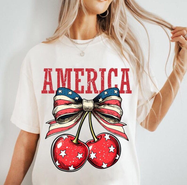 May include: White t-shirt with a red, white, and blue bow with stars and the word "AMERICA" printed on it. The bow is attached to two red cherries with white stars.