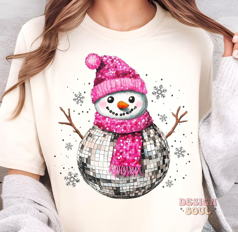 May include: A white t-shirt with a pink and silver snowman design. The snowman is wearing a pink sequined hat and scarf and has a disco ball for a body. The background is white with snowflakes.
