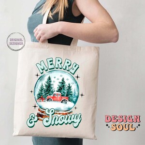May include: A white canvas tote bag featuring a red truck inside a snow globe. The text "Merry & Snowy" is printed in green on the bag. The words "Design Soul" are printed in pink and orange on the bottom right corner of the bag.