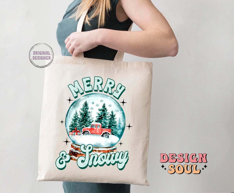 May include: A white canvas tote bag featuring a red truck inside a snow globe. The text "Merry & Snowy" is printed in green on the bag. The words "Design Soul" are printed in pink and orange on the bottom right corner of the bag.