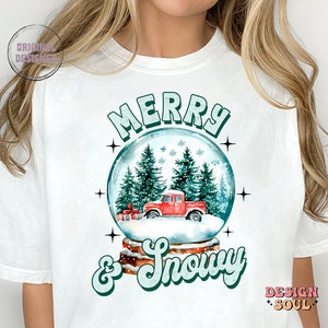 May include: White t-shirt featuring a green and white graphic of a snow globe with a red truck inside. The text "Merry & Snowy" is written below the snow globe.