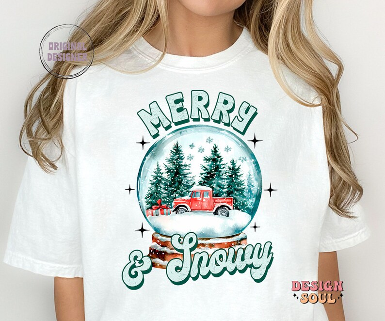 May include: White t-shirt featuring a green and white graphic of a snow globe with a red truck inside. The text "Merry & Snowy" is written below the snow globe.
