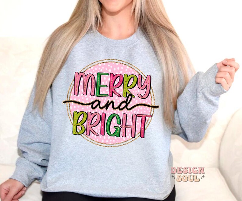 May include: A light gray sweatshirt with the words "MERRY and BRIGHT" in a colorful, playful font. The text is set against a pink polka-dot background within a gold circle. The sweatshirt has long sleeves.