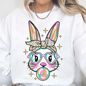 May include: White sweatshirt with a colorful cartoon bunny wearing heart-shaped sunglasses and a polka dot headband. The bunny is blowing a bubblegum bubble.