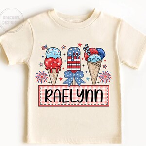 May include: A white t-shirt with a red, white, and blue ice cream cone design. The ice cream cones are decorated with stars and stripes. The text "Raelynn" is printed on a red and white checkered banner.