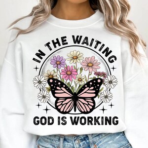 May include: White sweatshirt with a black and pink butterfly graphic and the text "In the waiting, God is working". The graphic also features a circle of colorful flowers.