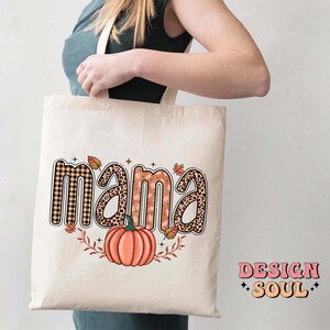 May include: A white tote bag with the word "mama" in brown, orange, and black patterned letters. The letters are decorated with fall-themed designs, including plaid, leopard print, and leaves. A small orange pumpkin is centered below the word "mama".