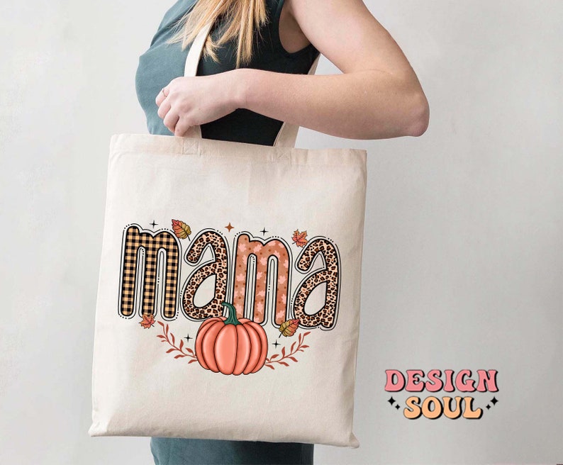 May include: A white tote bag with the word "mama" in brown, orange, and black patterned letters. The letters are decorated with fall-themed designs, including plaid, leopard print, and leaves. A small orange pumpkin is centered below the word "mama".