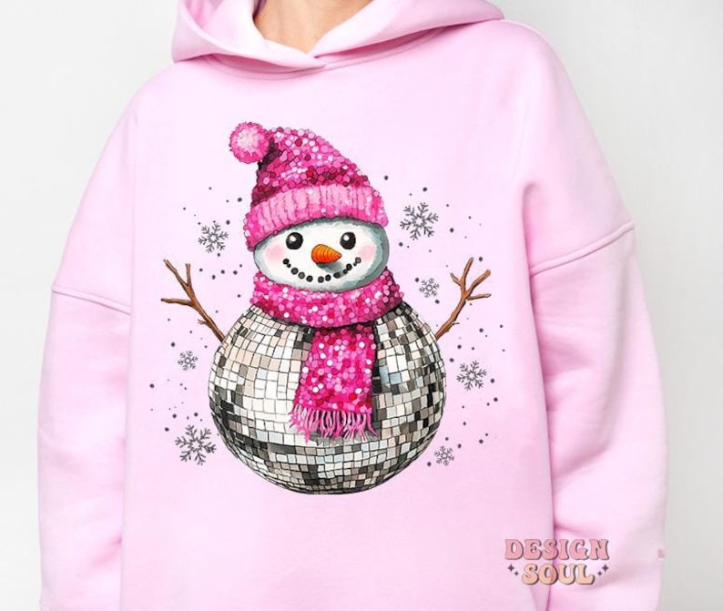 May include: Pink hoodie with a sparkly disco ball snowman wearing a pink hat and scarf. The text "DESIGN SOUL" is printed on the bottom right corner of the hoodie.