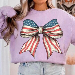Peut inclure: Un sweat-shirt lavande avec un grand graphique d'un n&oelig;ud rouge, blanc et bleu. Le n&oelig;ud est dessin&eacute; dans un style vintage et ressemble au drapeau am&eacute;ricain.