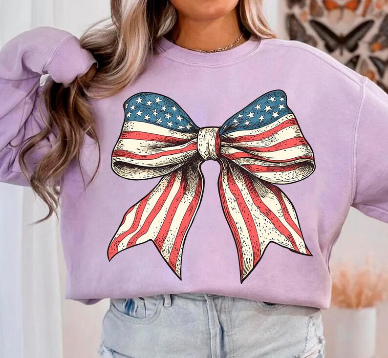 Peut inclure: Un sweat-shirt lavande avec un grand graphique d'un n&oelig;ud rouge, blanc et bleu. Le n&oelig;ud est dessin&eacute; dans un style vintage et ressemble au drapeau am&eacute;ricain.